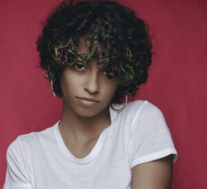 Artistic portrait of a woman in a white shirt with colorful hair on a red backdrop.